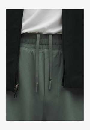 Primer plano de pantalones grises con cintura elástica y cordones, combinados con una camisa blanca y una chaqueta negra con cremalleras.