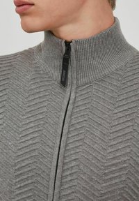 Grå sweater med høj krave, der har en zigzag ribbet tekstur og en lynlås lukning. Omfatter en sort lynlåsbund med branding.