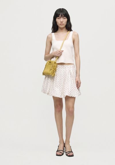 Une femme porte un haut blanc sans manches, une jupe à motifs, des sandales noires et un sac bandoulière jaune à franges, sur un fond clair uni.