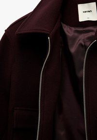 Bordeaux wollen jas met een ritssluiting, brede kraag en een enkele zak. De binnenkant heeft een gladde, donkere stofvoering.