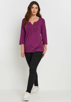 Paarse langwerpige top met een V-hals en knoopdetails, met plooien op de borst. Gekozen in combinatie met zwarte leggings en witte sneakers.