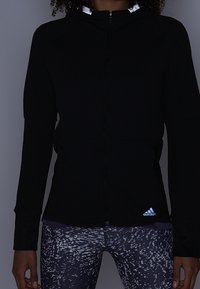 Veste noire zippée avec une surface texturée et des accents réfléchissants, associée à un legging à motifs. Le logo Adidas est visible sur la veste.