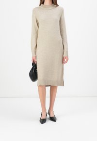 Abito a maglia beige con colletto alto e maniche lunghe, fessure laterali all'orlo, abbinato a una borsa nera e tacchi appuntiti neri.