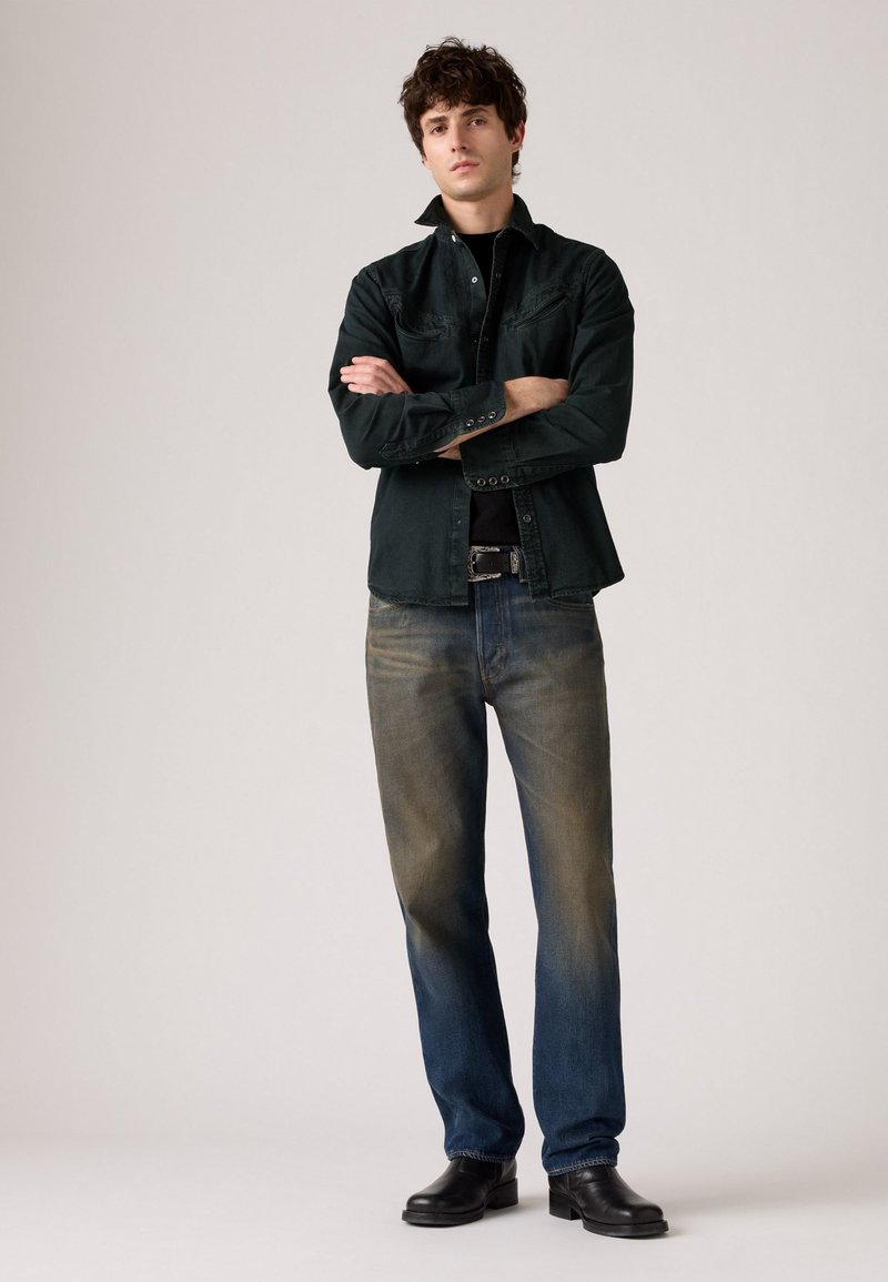 Veste en denim vert foncé, t-shirt noir en dessous, jeans bleus délavés et bottes en cuir noires. Le mannequin se tient avec les bras croisés, face à l'objectif.