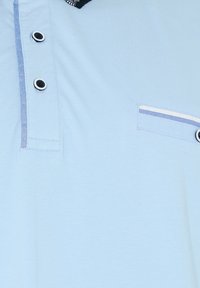 Chemise bleu clair avec deux boutons noirs et blancs sur la patte de boutonnage et un petit bord de poche avec un liseré bleu et blanc.