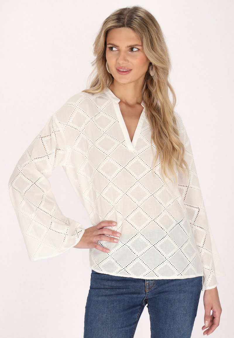 Weiße Bluse mit langen Ärmeln und diamantförmigen Ausschnitten sowie einem V-Ausschnitt. Der Stoff sieht leicht und halbtransparent aus. Zusammen mit Jeans getragen.