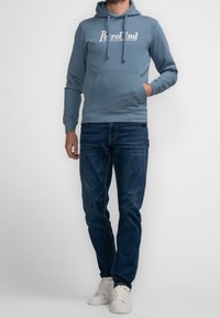 Světle modrá mikina s bílým logem, připojenou kapucí a klokaní kapsou; kombinovaná s tmavě modrými džíny a bílými teniskami.