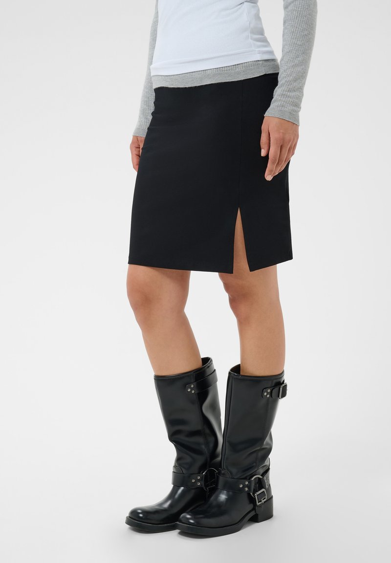 Personne portant une jupe noire fendue à l'avant, un haut gris à manches longues et des bottes en cuir noires mi-mollet avec des boucles.