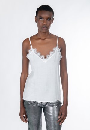 Femme portant un caraco blanc orné de dentelle et un pantalon argenté brillant, debout devant un fond blanc uni.