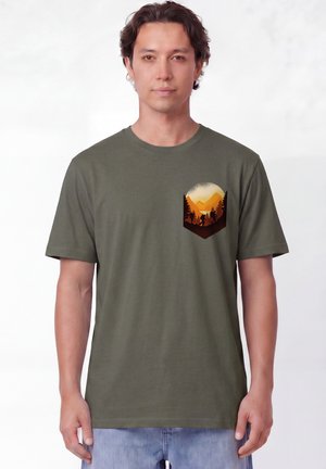 Homme portant un t-shirt vert olive avec un motif sur la poche d’un coucher de soleil sur des montagnes et des arbres avec deux randonneurs qui marchent.