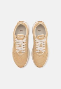 Beige semsket sneakers med hvite snøringer, rund tå, perforerte detaljer og ribbet brune gummisåler, med preget 'BOSS' logo på tungen.