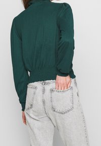 Turkos texturerad blus med samlad krage och puffärmar, instoppad i ljusgrå högmidjade jeans med bakfickor.