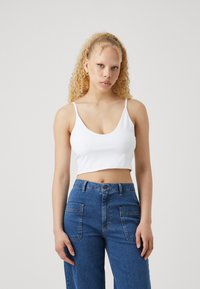 Valge puuvillane crop top õhukeste rihmadega ja laia kaelusega, kaasas kõrge vöökohaga sinised teksapüksid, millel on suured tagataskud.