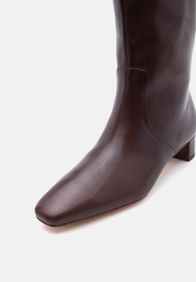 Σκούρο καφέ δερμάτινη μποτάκι μέχρι τη μέση της γάμπας με τετράγωνη μύτη και χαμηλό χοντρό τακούνι, εμφανίζεται από κοντινή γωνία σε ανοιχτό φόντο.