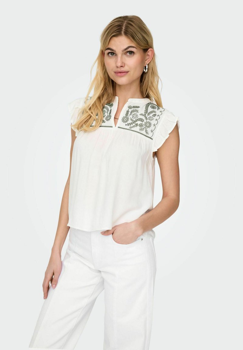 Jeune femme aux longs cheveux blonds portant un chemisier blanc sans manches avec broderie florale verte et pantalon blanc, posant avec une main dans la poche.
