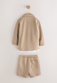 Chemise beige à manches longues avec col et short élastique assorti à la taille suspendu à un crochet en bois contre un mur clair.