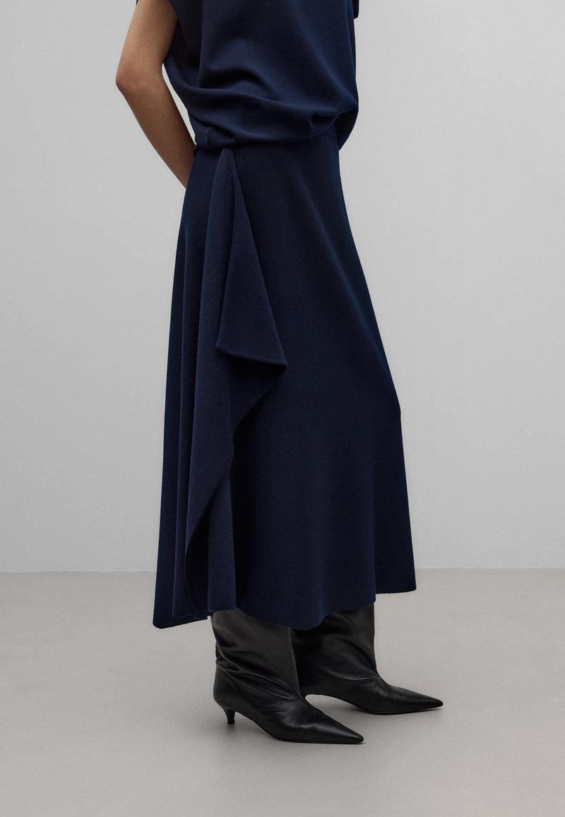 Femme portant une jupe mi-longue bleu marine avec un détail drapé asymétrique, associée à des bottines à talons noires à bout pointu, sur un fond uni.