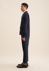 Giacca di lino navy con una tasca sul petto, abbinata a pantaloni coordinati. Scarpe in pelle nera completano il look. Design minimalista, vestibilità su misura.