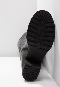 Botte en cuir noir avec une semelle en caoutchouc épaisse, faisant face à l'appareil photo, reposant contre un mur blanc sur une surface blanche.