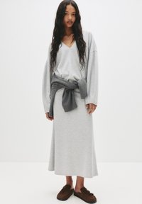 Sweatshirt gris clair avec col en V, manches longues et un détail de taille noué gris foncé, associé à une jupe longue assortie et des chaussures à enfiler marron.