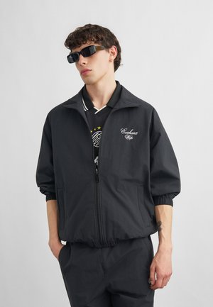 Junger Mann trägt eine schwarze Jacke mit Reißverschluss und dem "Carhartt Wip"-Logo, schwarze Hose, dunkle Sonnenbrille und ein schwarzes Hemd mit weißem Kragen.