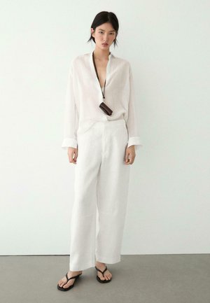 Woman wearing loose white shirt and wide white pants, black flip-flops, and a brown accessory hanging from her neck against plain background.