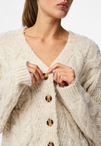 Μπεζ πλεκτή ζακέτα με V-λαιμόκοψη, που διαθέτει μεγάλα καφέ κουμπιά, περίτεχνα σχέδια με πλέξη καλωδίου και ριμπ μανσέτες για υφή και λεπτομέρεια.
