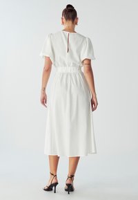 Biała midi sukienka z krótkimi bufiastymi rękawami, marszczoną talią i kluczowym detalem na plecach. Noszona z czarnymi sandałami na obcasie o gładkiej fakturze.