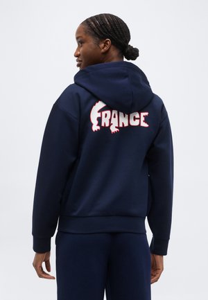 Žena s opletenými vlasmi, ktorá nosí tmavomodrú mikinu s kapucňou s nápisom "FRANCÚZSKO" v bielych a červených písmenách na chrbte, stojaca pred jednoduchým pozadím.