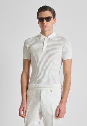 Uomo che indossa una polo bianca a maniche corte a coste, pantaloni bianchi e occhiali da sole scuri, in piedi davanti a uno sfondo grigio chiaro uniforme.