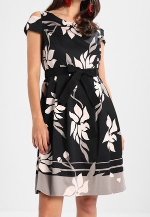 Robe noire mi-longue avec de grands motifs floraux beiges, manches épaules dénudées et ceinture nouée à la taille.