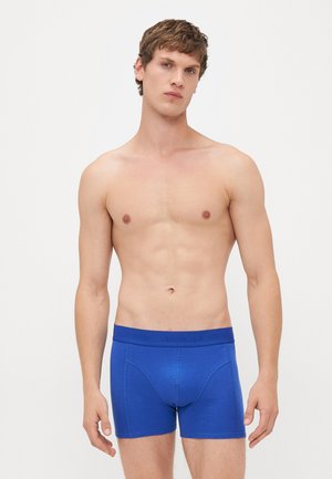 Junger Mann mit kurzen braunen Haaren, der royale blaue Jack & Jones Herrenunterwäsche trägt, vor einem schlichten weißen Hintergrund stehend.