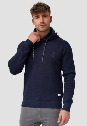 Navy blå hettegenser med teksturert strikk, justerbar snorhette og en subtil logo på brystet. Ribbestrikkede mansjetter og nederkant.