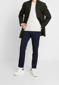 Blazer long vert foncé, pull gris clair, pantalon slim marine, et baskets blanches avec des accents marines. Tenue décontractée et polyvalente.
