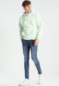 Hellgrüner Hoodie mit einer Kängurutasche, Kapuze mit Kordelzug und gerippten Bündchen, kombiniert mit blauen engen Jeans und weißen Sneakern.