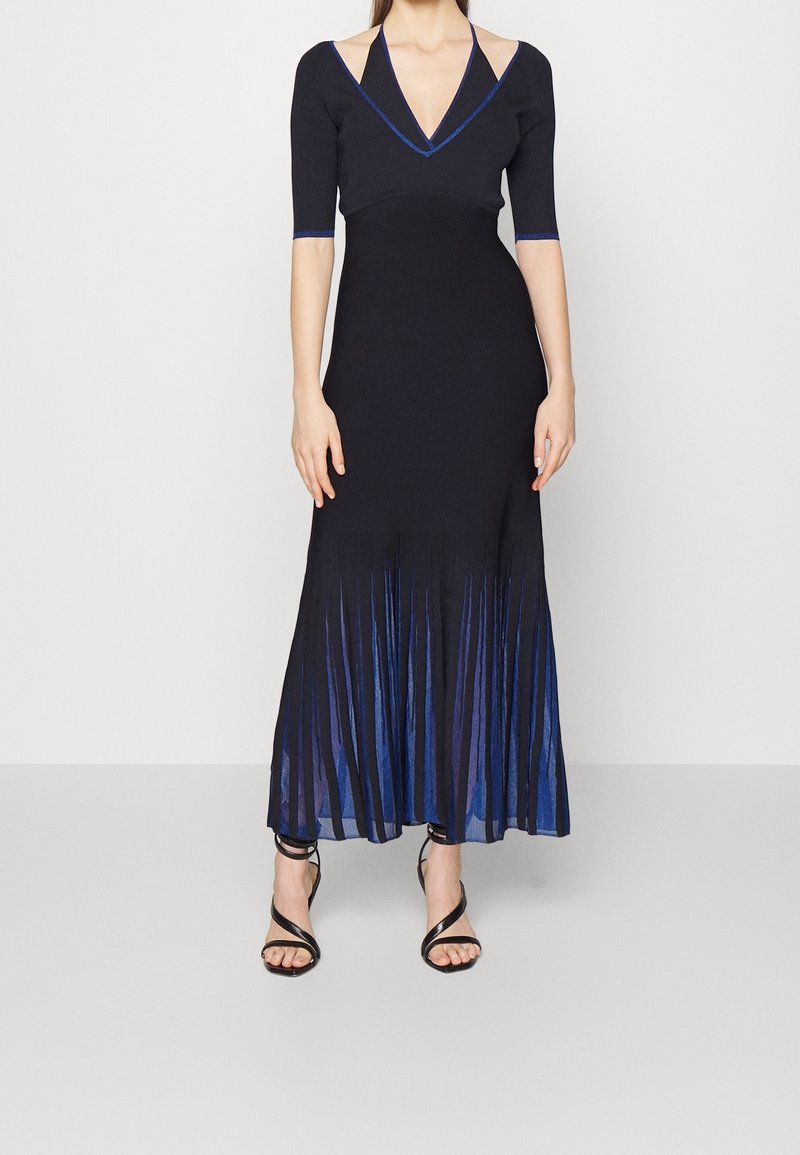 Femme portant une robe noire et bleue à col en V avec manches mi-longues, découpes aux épaules, ourlet plissé et sandales noires à talons hauts avec brides.