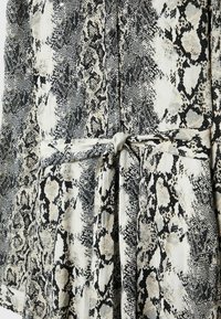 Gros plan d'une robe-chemise à imprimé serpent boutonnée avec une ceinture en tissu nouée à la taille dans des tons noir, blanc et gris.