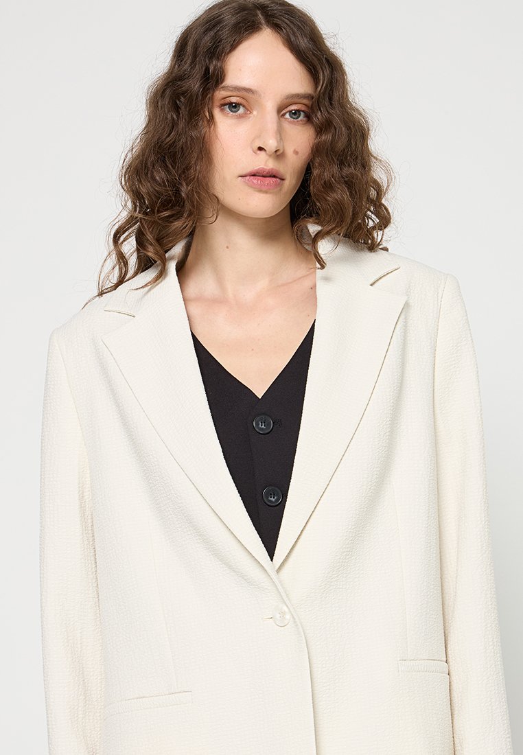 Weißer strukturierter Blazer mit Reverskragen, Ein-Knopf-Verschluss und zwei Fronttaschen, über einem schwarzen Oberteil mit Knöpfen getragen.