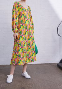 Gul klänning med blommönster och långa ärmar, med nyanser av rosa, grönt och lila. Kompletteras med vita sneakers och en grön väska.