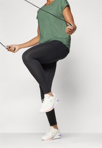 Leggings neri, maglietta verde a maniche corte e sneaker bianche con dettagli viola. Atleta che usa una corda per saltare, con una gamba sollevata.