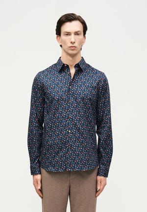 Άνδρας με σκούρα μαλλιά, φοράει μπλε σκούρο πουκάμισο με μακριά μανίκια και μικρά κόκκινα και λευκά γεωμετρικά σχέδια, σε συνδυασμό με μπεζ παντελόνι.