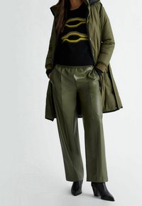 Femme portant un pantalon large vert olive, des bottes à talons noires, un pull noir avec un motif jaune, et un long manteau matelassé vert olive.