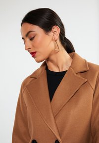 Cappotto di lana marrone con colletto a scollo e bottoni neri. Top nero sotto. Orecchini a triangolo dorati. Texture liscia, vista di profilo.