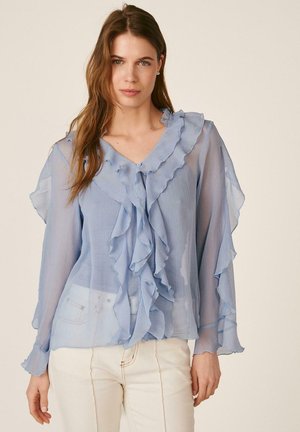 Mujer con blusa azul claro translúcida con volantes en el frente y mangas largas, combinada con pantalones de color claro, de pie frente a un fondo liso.