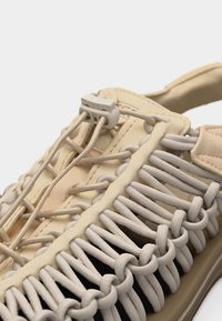 Beeži sandaali lähivõte, kus on näha sõlmitud elastsed nöörid ja ülasideme reguleeritav plastist lukustus.