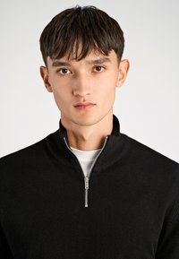 Schwarzer Zip-Hals-Pullover mit geripptem Kragen, aus weichem Strickmaterial, mit einem silbernen Reißverschluss und über einem weißen Hemd getragen.