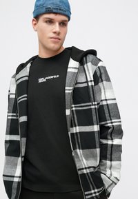 Jachetă din flanel în carouri alb-negru, purtată peste o cămașă neagră cu logo-ul "KARL LAGERFELD JEANS". Jacheta are glugă, combinând texturi moi.