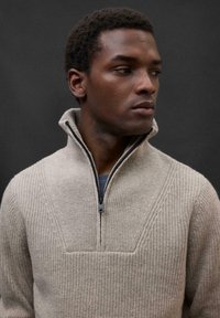 Beiger Strickpullover mit einem halben Reißverschlusskragen, der über eine gerippte Textur und ein geometrisches Muster auf der Brust verfügt. Glatte Stoffoberfläche.