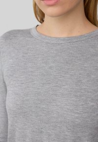Kobieta w jasnoszarej, długouchwowej koszulce typu crew neck o widocznej strukturze, skróconej poniżej nosa.
