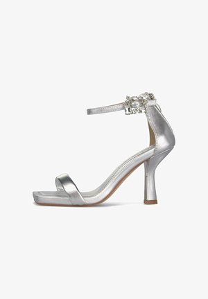 Srebrny sandał na wysokim obcasie z paskiem wokół kostki ozdobionym dekoracyjną sprzączką z kryształków, odkrytym noskiem i kwadratową podeszwą.